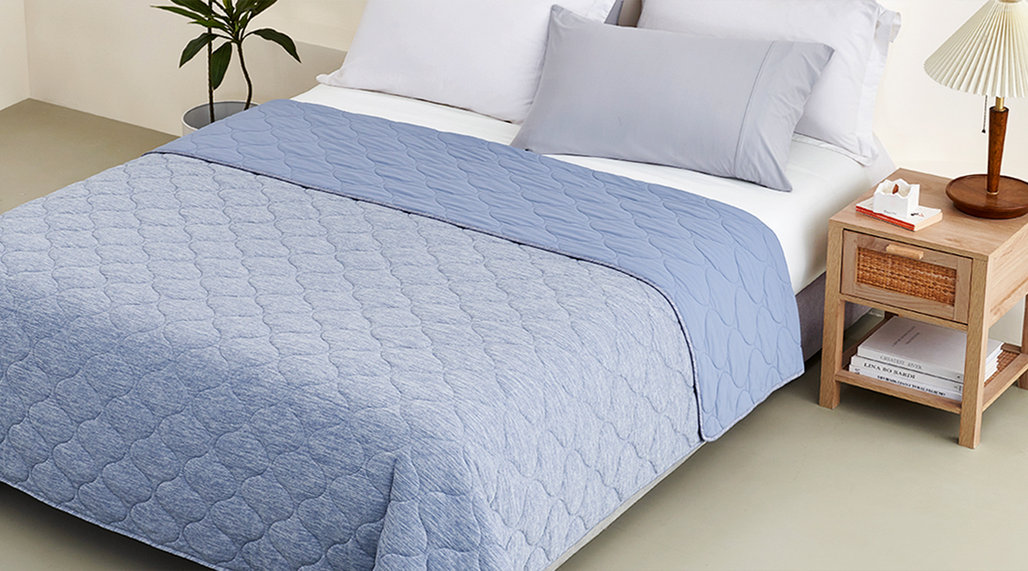A light blue quilted bedspread covers a bed with white sheets and a light gray pillow, next to a wooden nightstand.