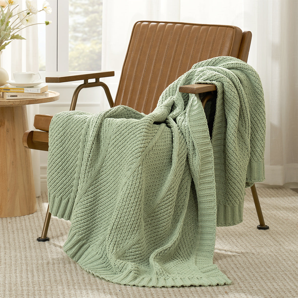 Ivory knitted blanket draped over a sage green armchair in a room with a table and window. #color_Sage Green