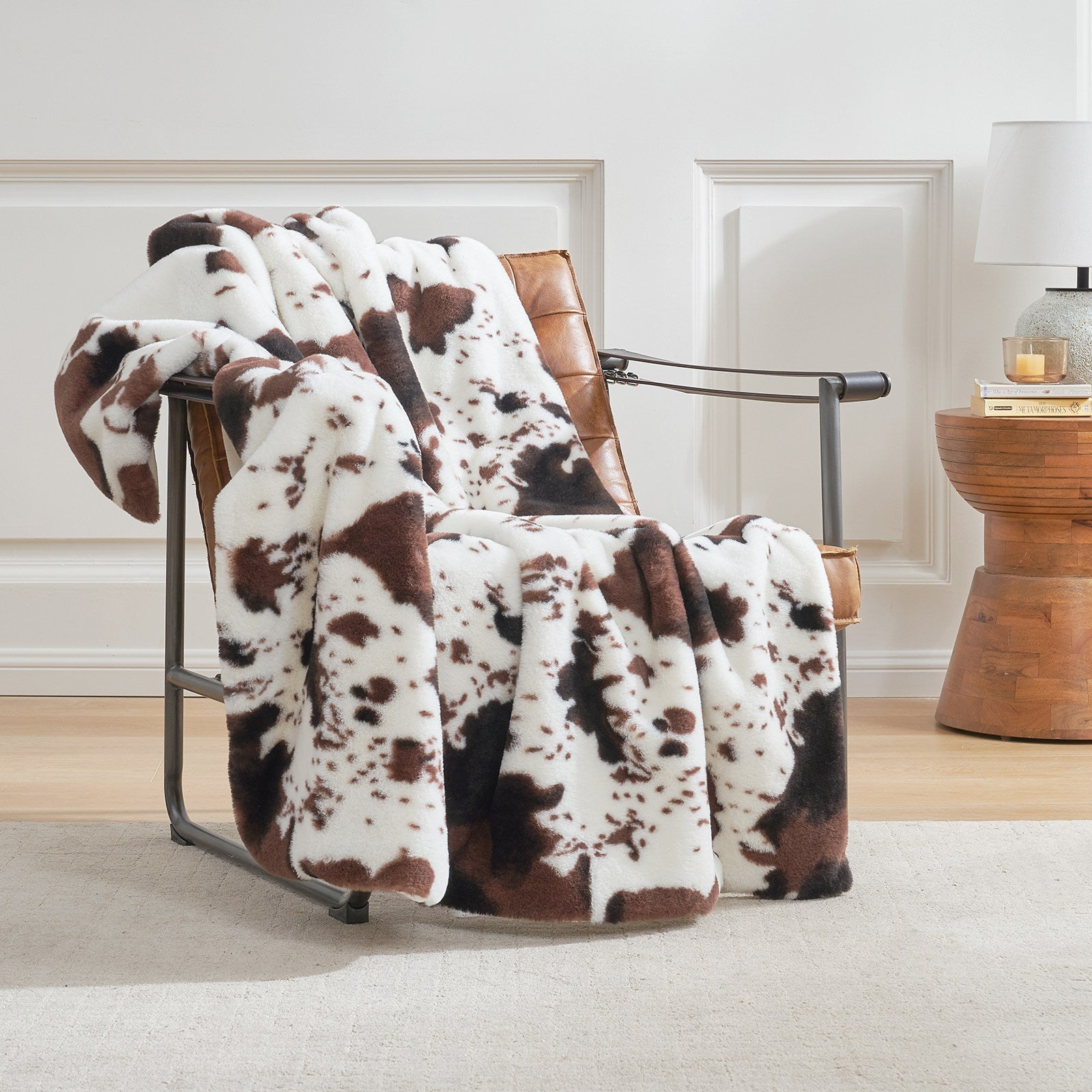 Cow print faux fur blanket draped over leather chair. Soft, cozy cow print blanket for home decor. #color_Black/Coffee