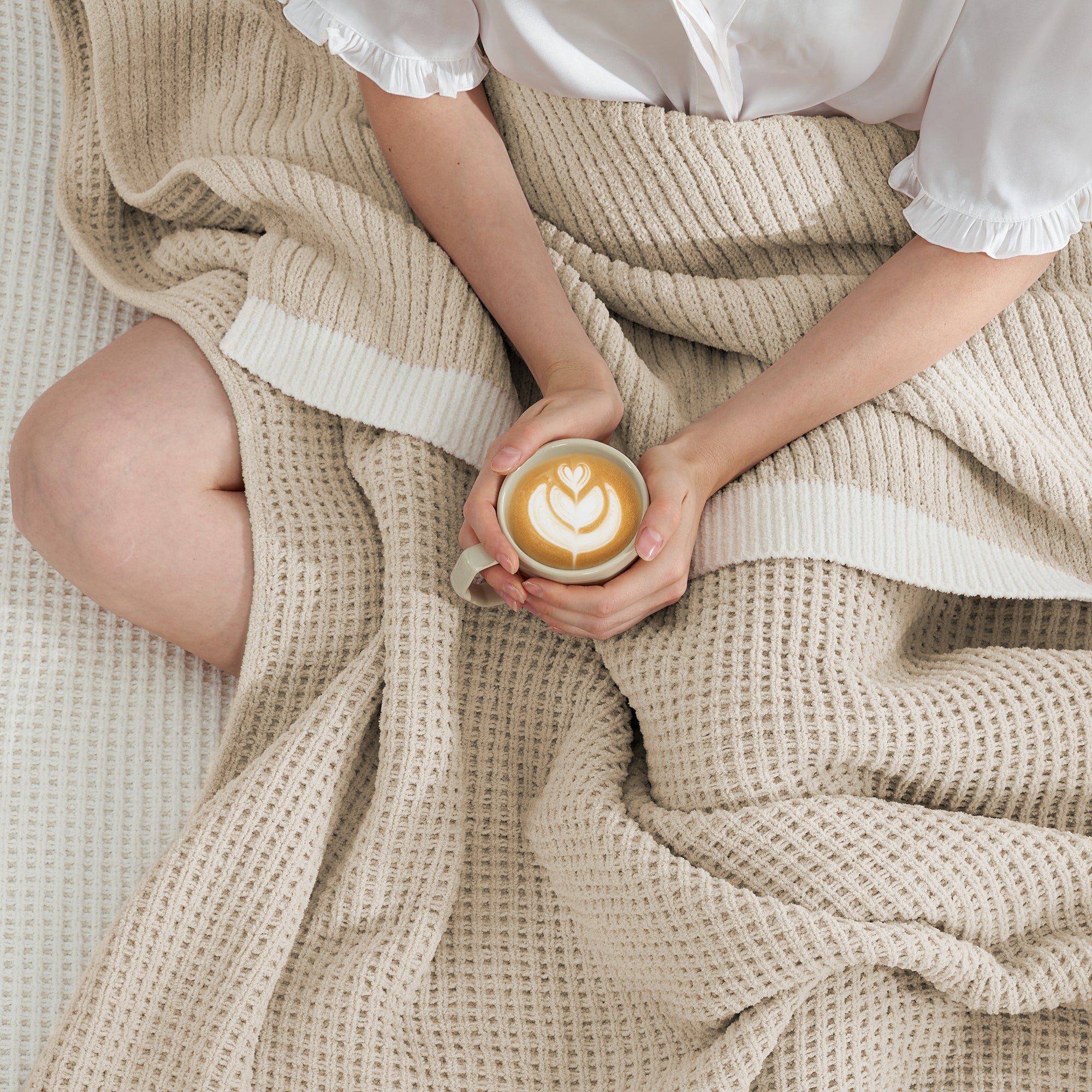 Woman wrapped in a Chenille Waffle Knit Throw Blanket in beige, enjoying a cup of coffee on a sofa. #color_Beige