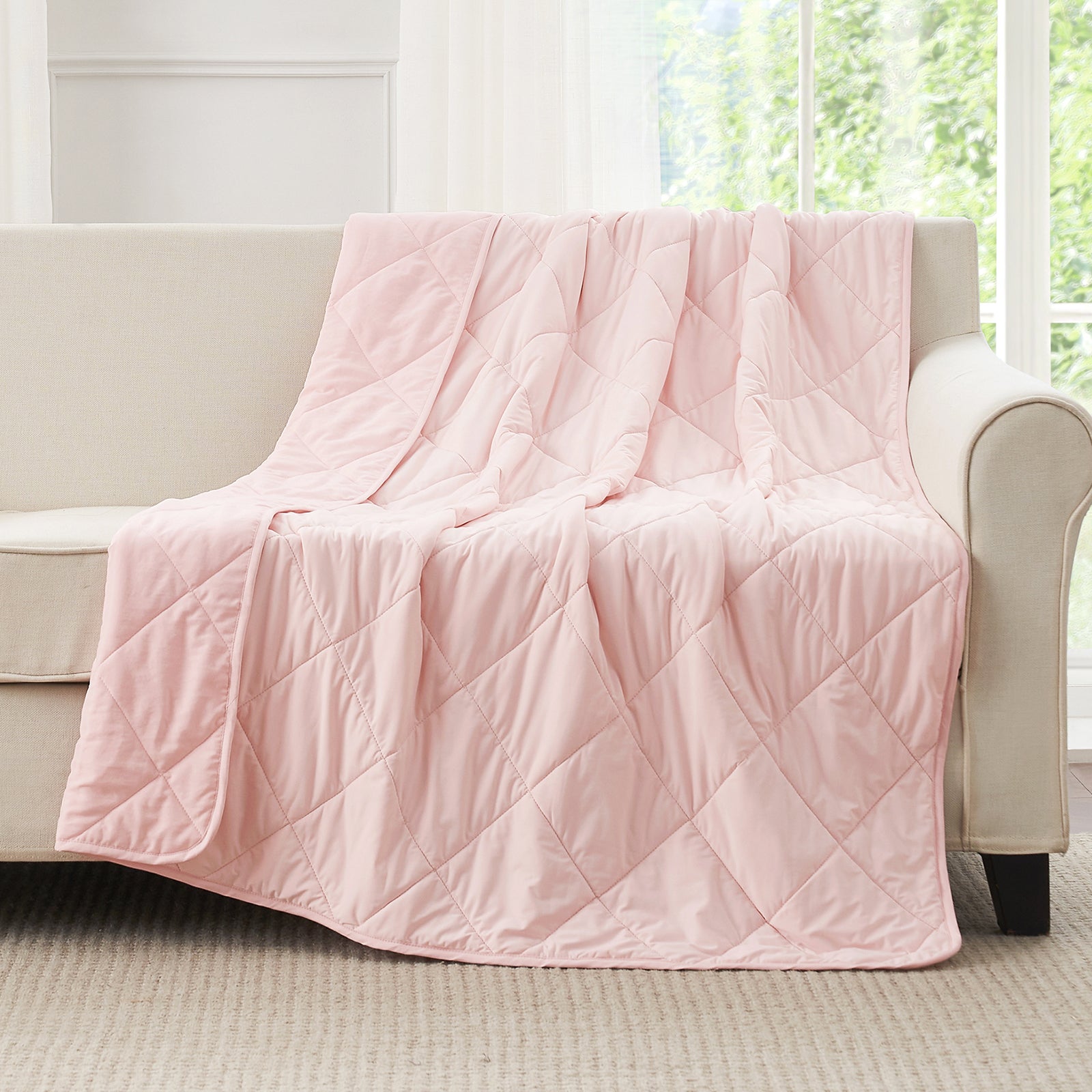 Cool pink quilted blanket draped over a beige sofa in a bright room with large windows. #color_Cool Pink
