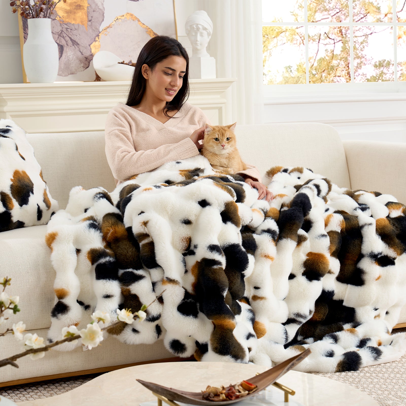 Woman sitting on a couch with a cat, wrapped in a cow print blanket, in a cozy living room. #Color_Cow