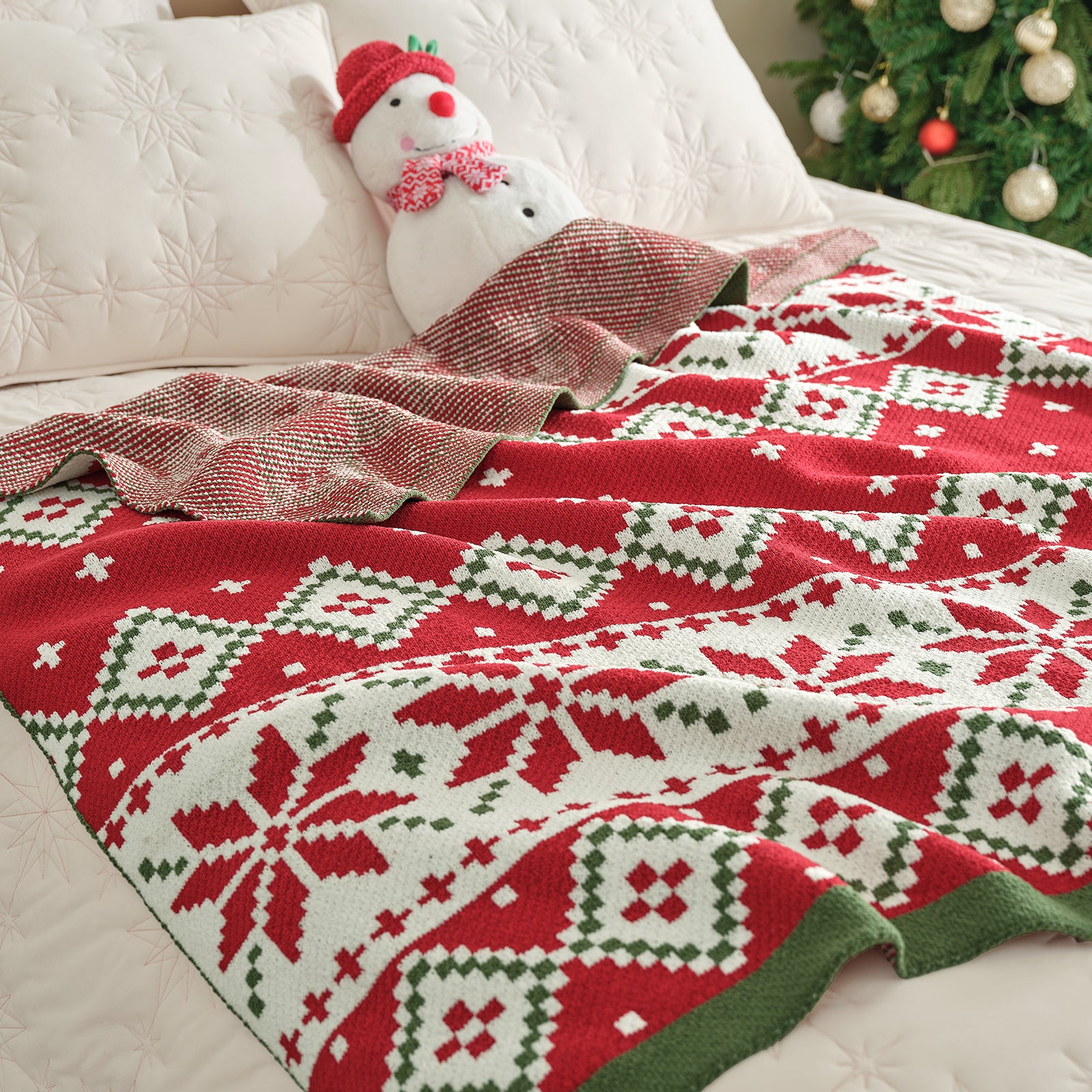 Red and green patterned blanket draped over a bed with a snowman pillow and Christmas tree in the background. #color_Fair Isle