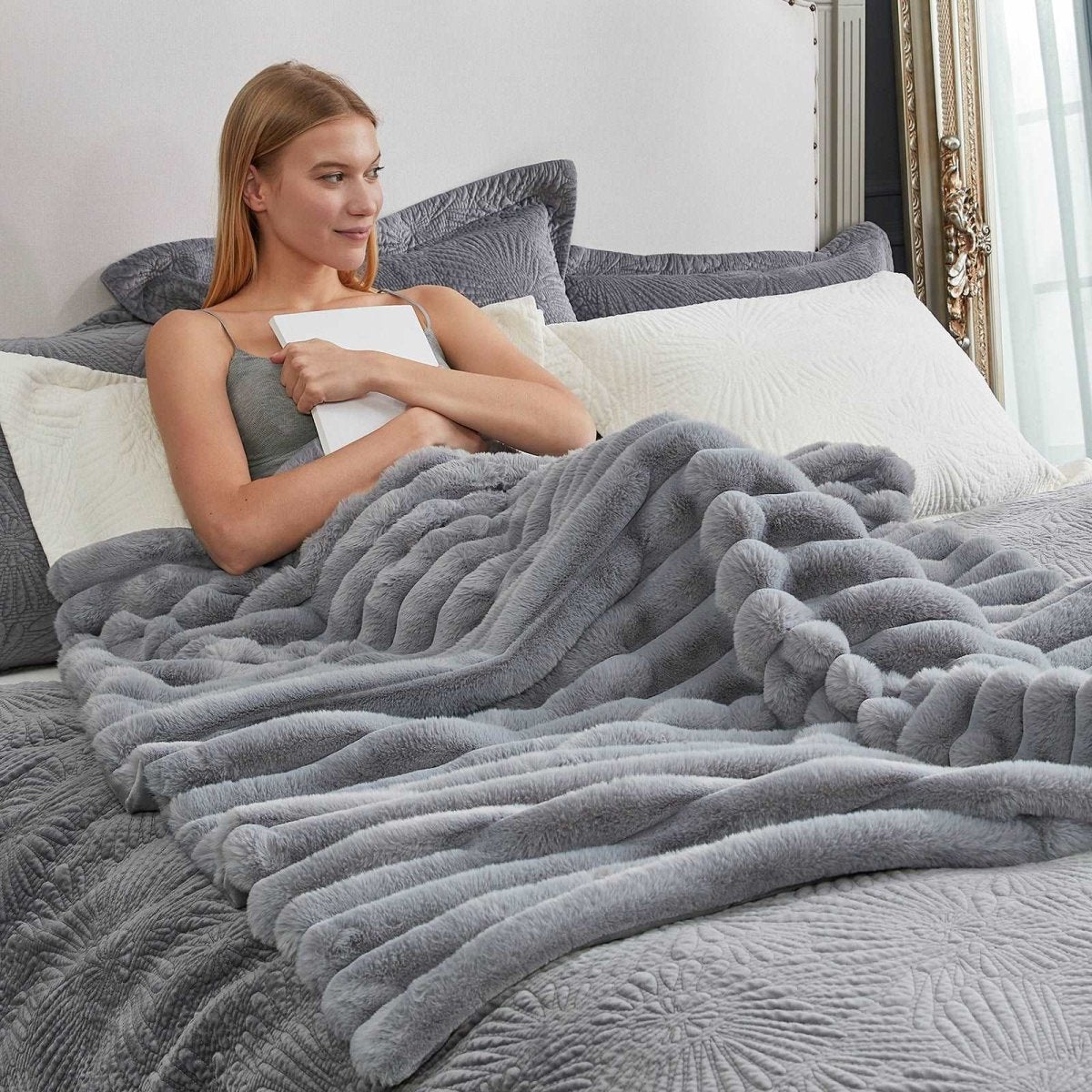 Woman lying on bed with Cozy Bliss grey faux fur blanket, cozy bedding and soft blanket. #color_Grey