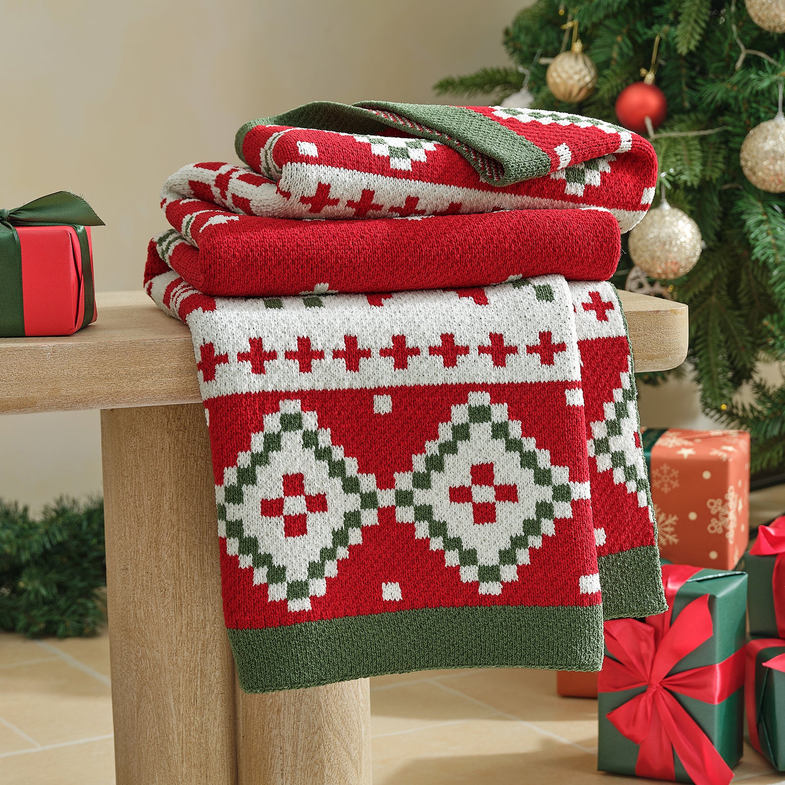 Stack of patterned red and green blankets on a wooden surface with Christmas decorations in the background. #color_Fair Isle
