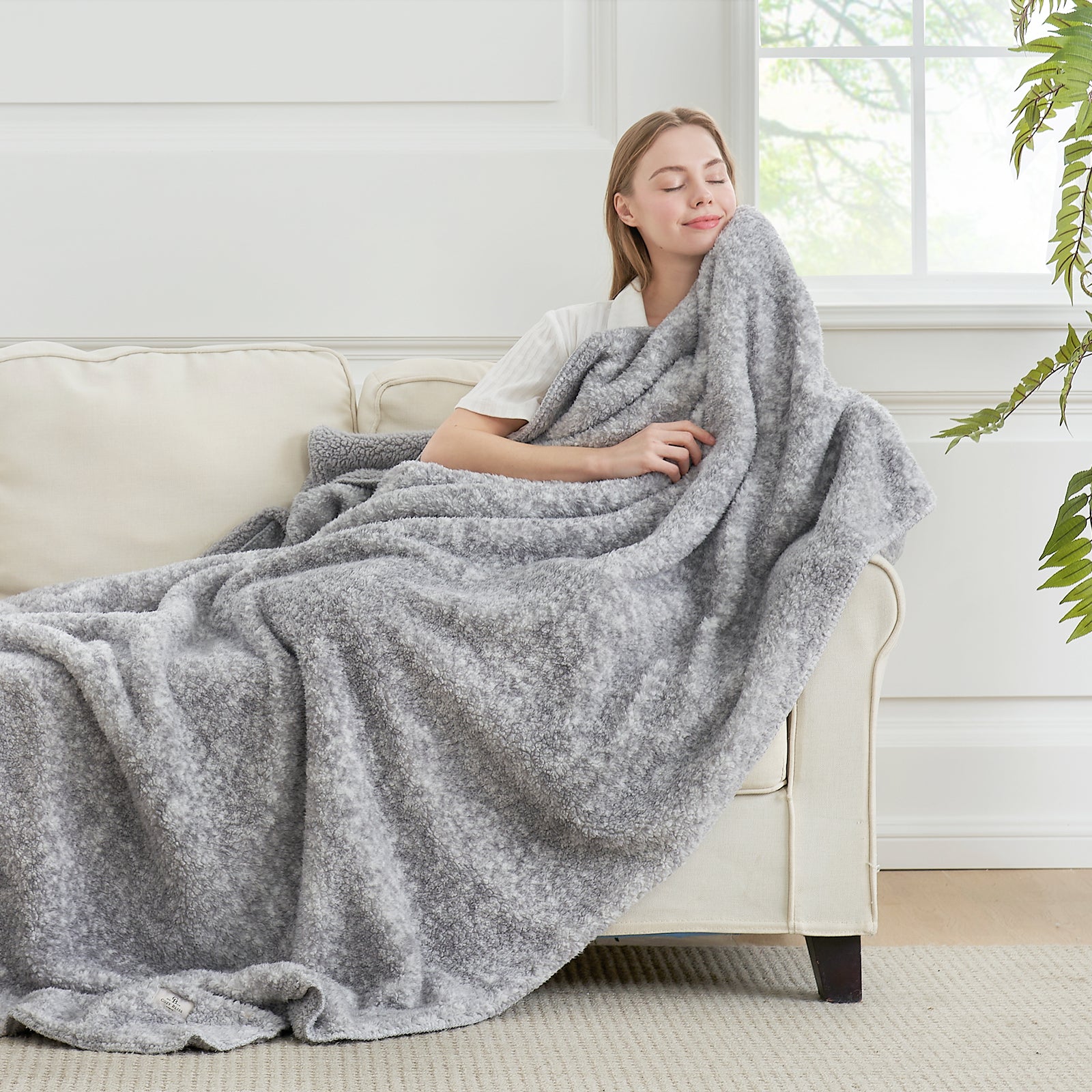 Woman relaxing with soft gray Sherpa fleece throw blanket on a sofa. Cozy blanket for comfort. #color_ Mottled Grey