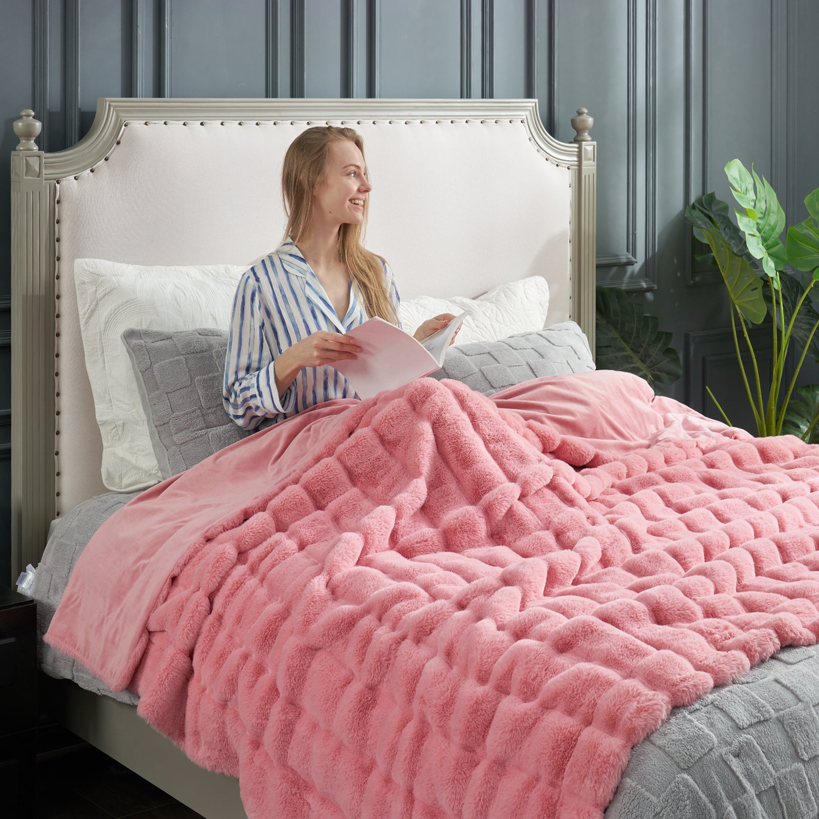 Woman sitting on a bed with pink comforter and gray pillows, reading a book. #color_Pink