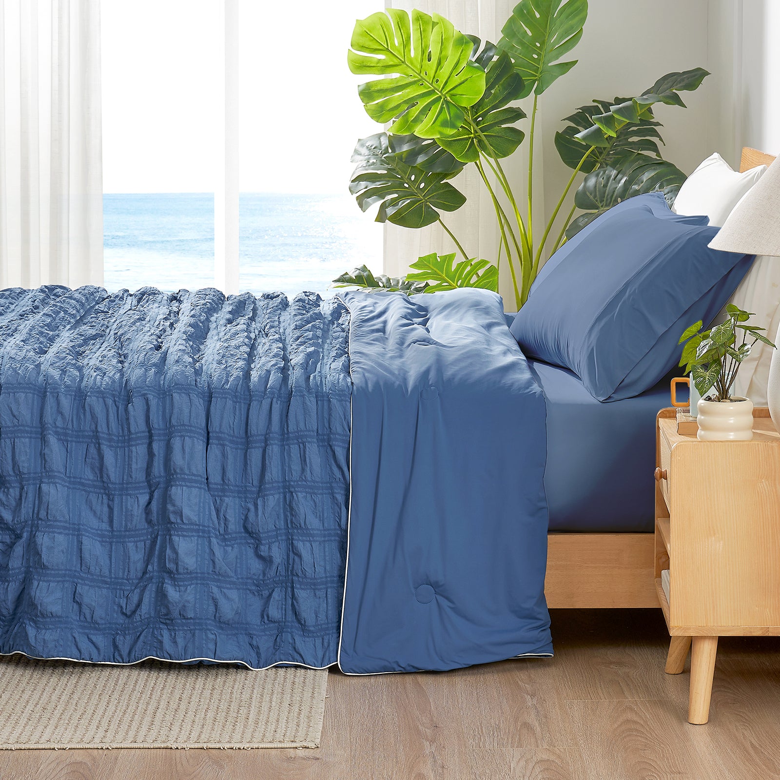 Blue bedding set on a bed with a wooden nightstand and plants in a room with a window view. #color_Denim Blue