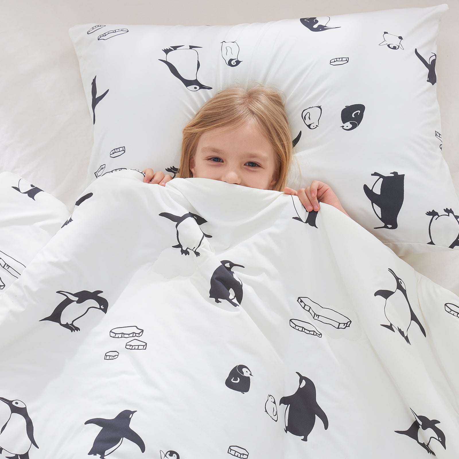 Child under a white comforter with penguin patterns, lying on a bed with matching pillowcases.#color_Antarctic Penguin