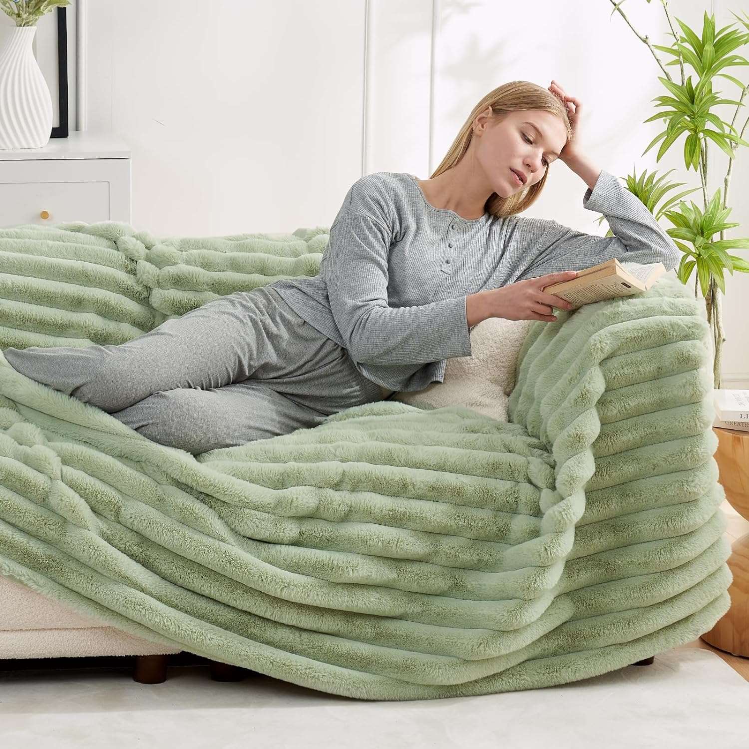 Woman relaxing under a sage green striped faux fur blanket on a sofa. Cozy bedding. #color_Sage Green