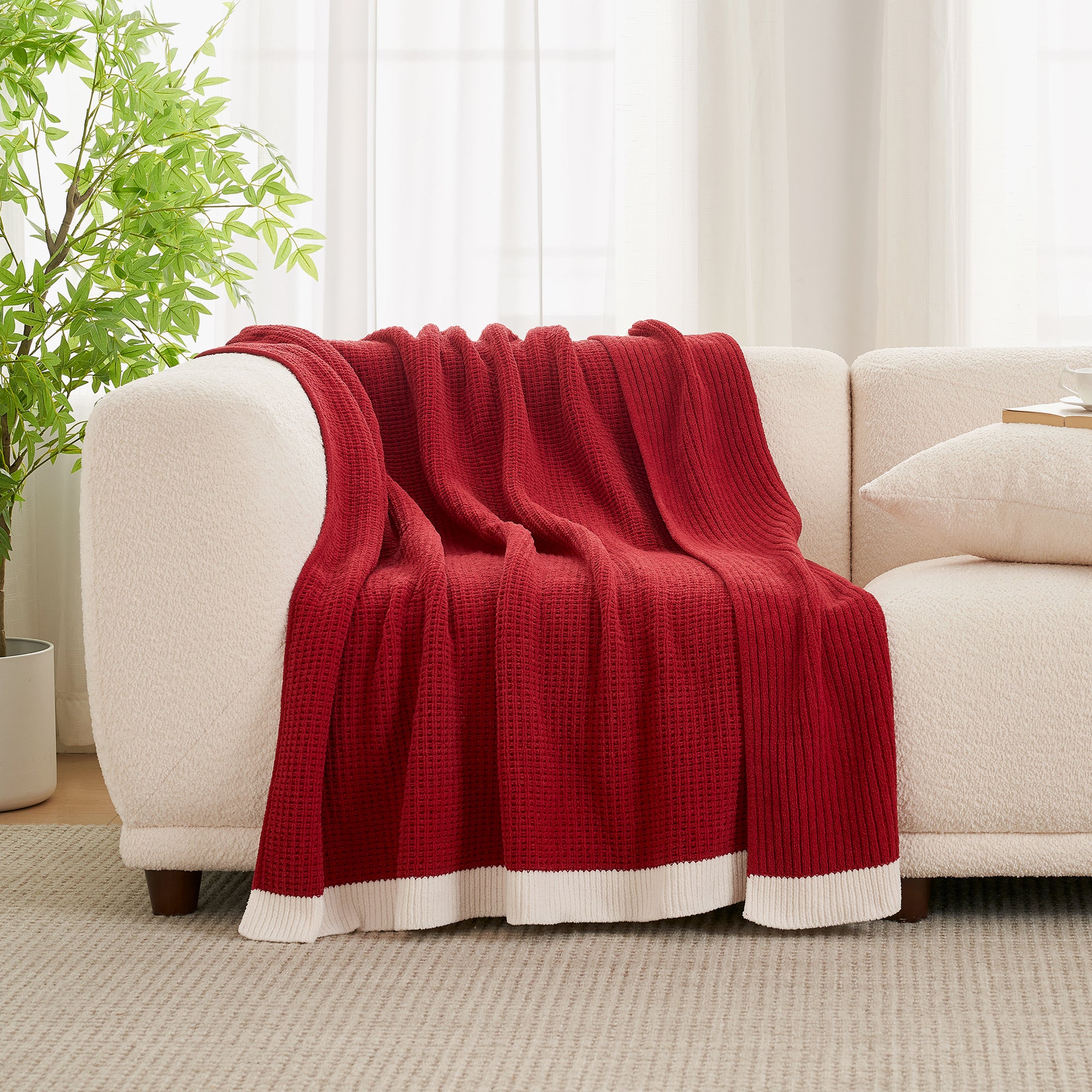 Red blanket draped over a beige sofa in a living room setting with a plant and curtains. #color_Red
