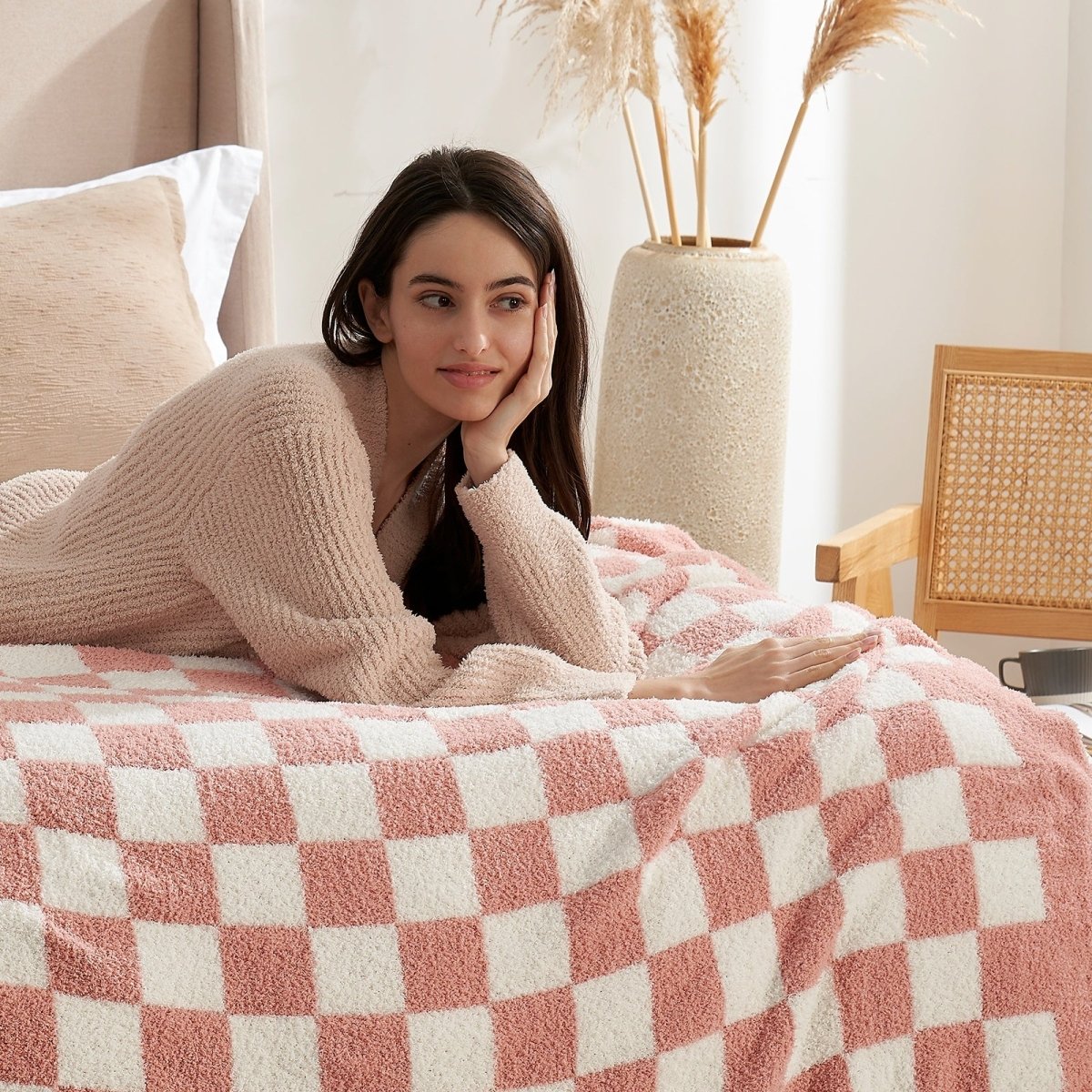 Woman lying on a bed with a Checkered Feather Yarn Knit Throw Blanket in Pink in a cozy living room setting. #color_Pink White