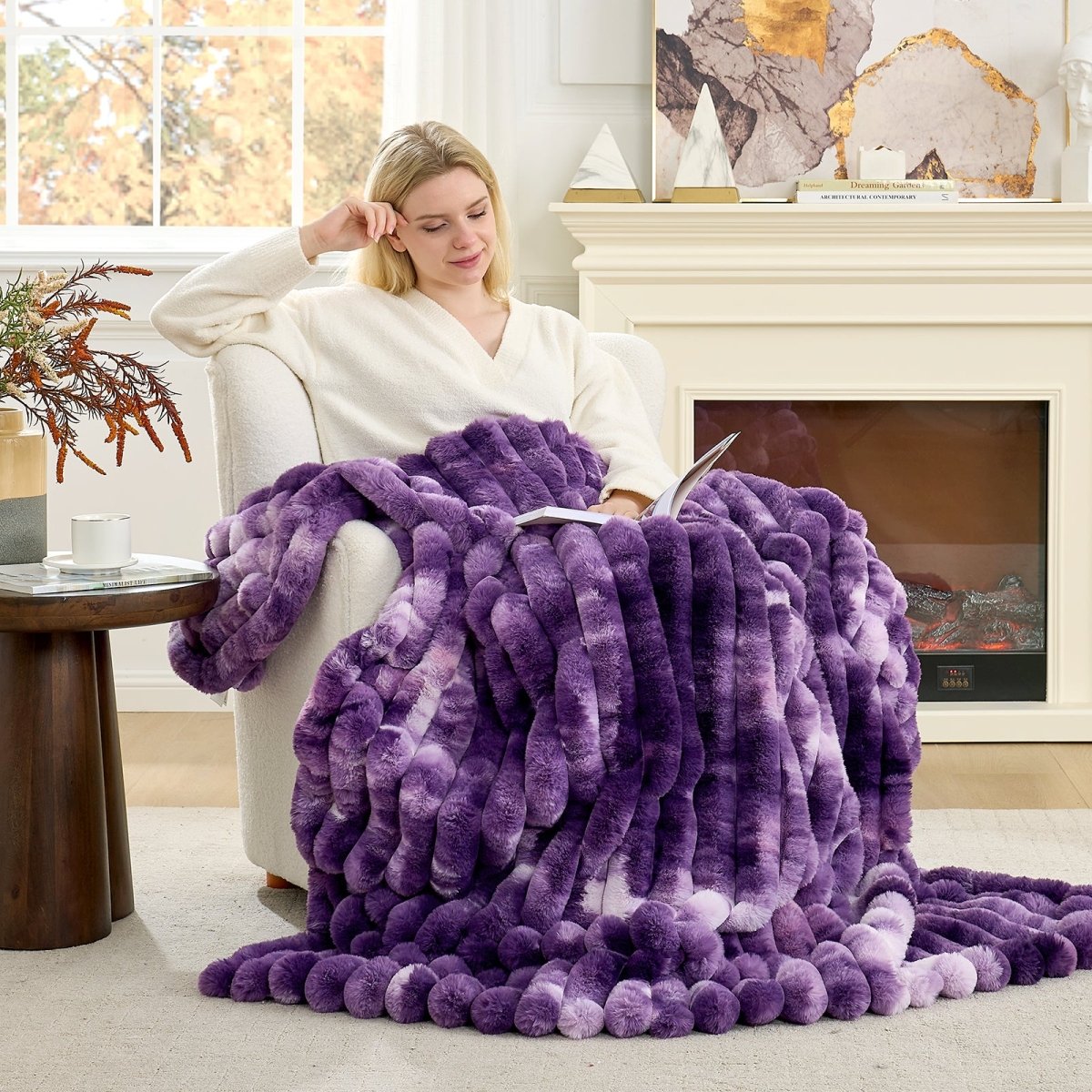 Woman sitting on a couch with a purple blanket, reading a book in a cozy living room. #color_Tie-dye Purple