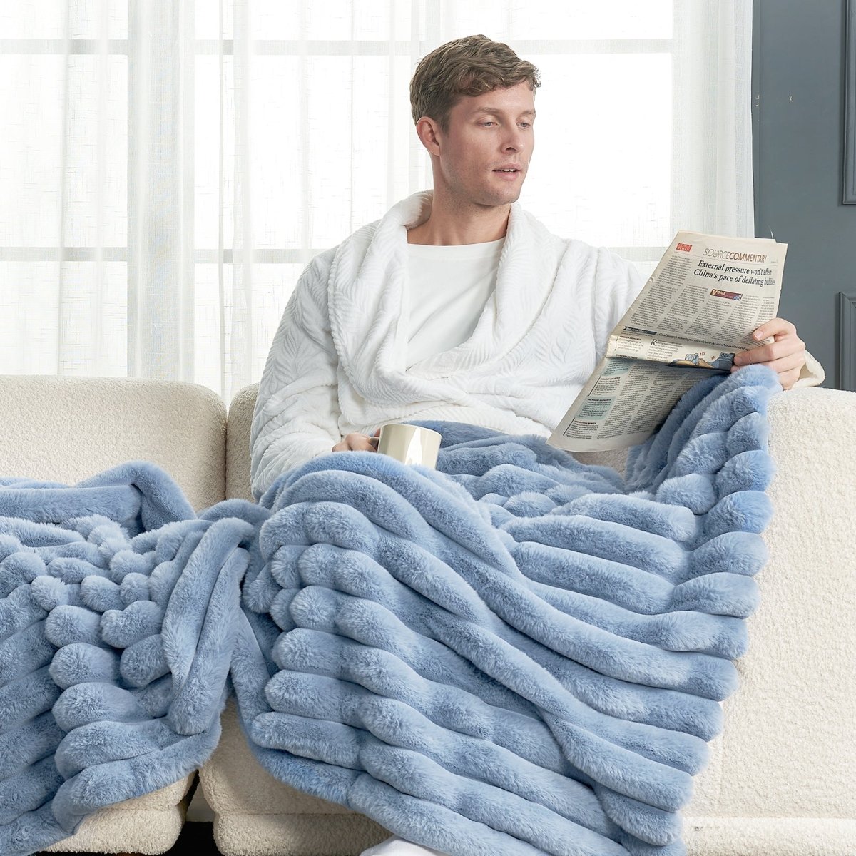 Man sitting on a couch with a light blue striped faux fur throw blanket draped over him, reading a newspaper. #color_Misty Blue