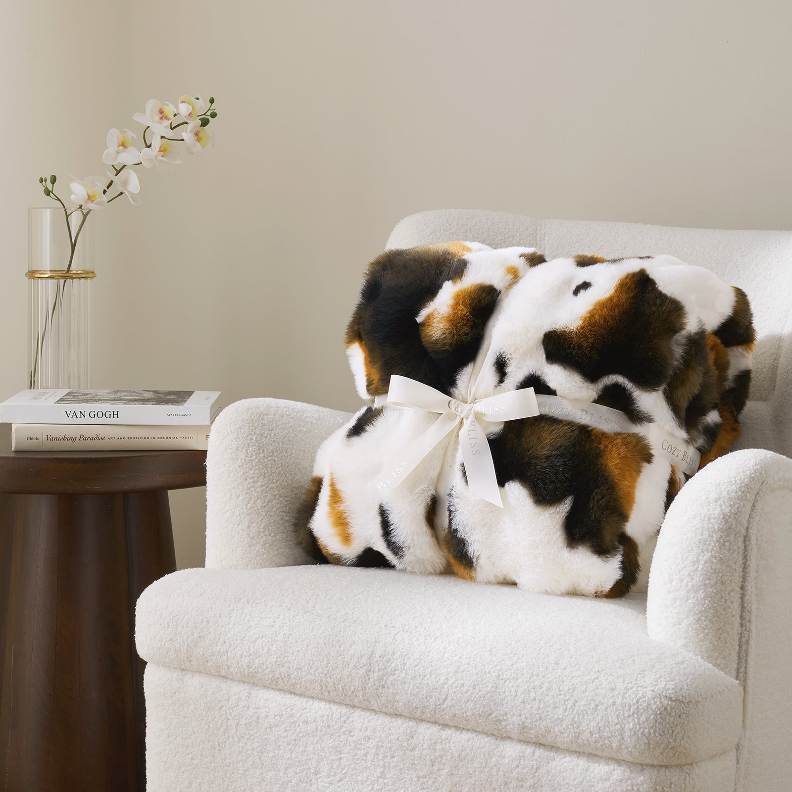 Fluffy cow print blanket with a bow on a white armchair, next to a small table with books and a vase. #Color_Cow