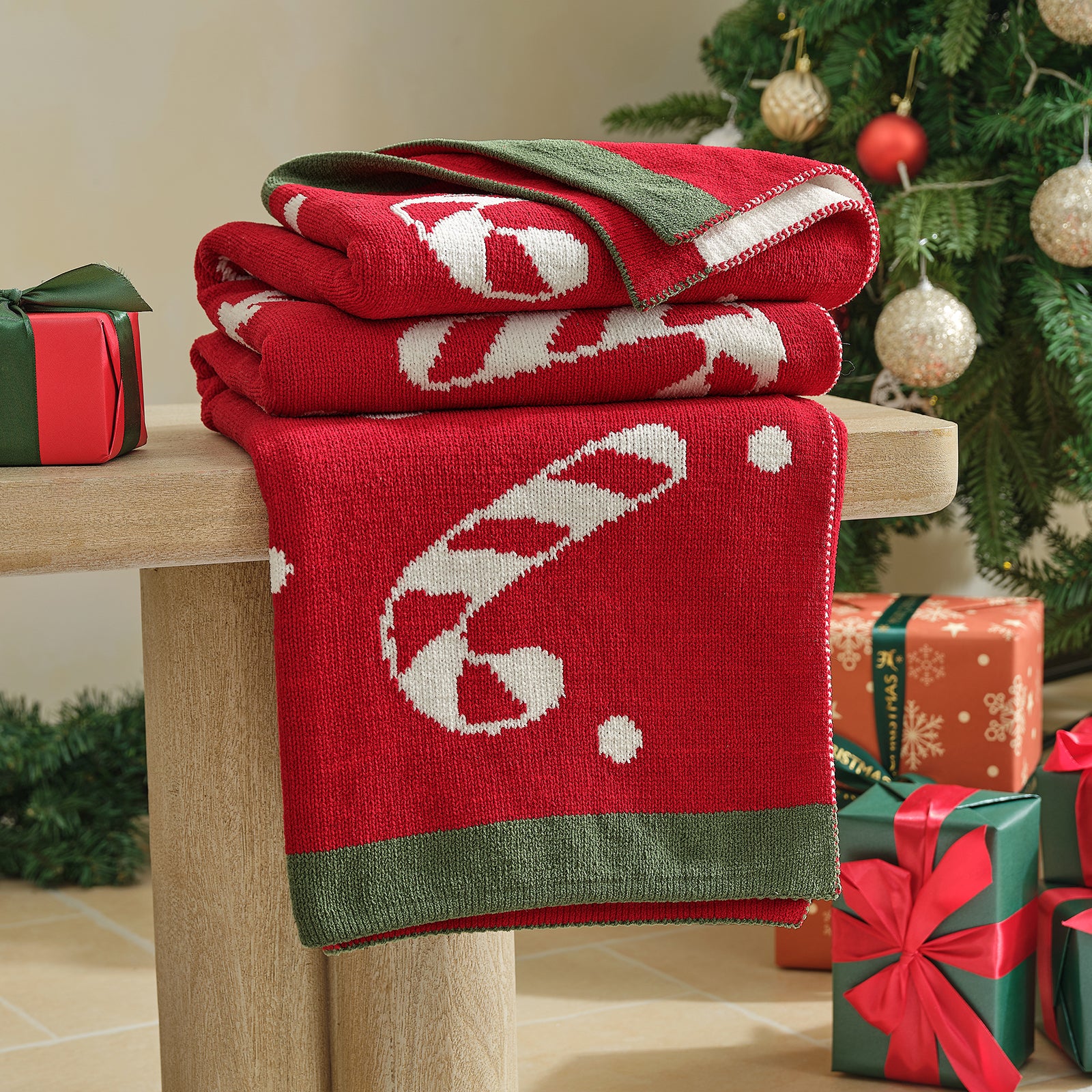 Stack of red Christmas-themed blankets on a wooden surface with a decorated tree in the background. #color_Candy Cane