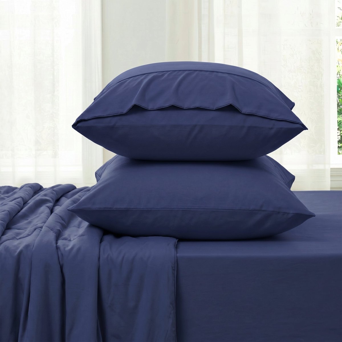 Blue bedding set with two pillowcases and fitted sheet against a white curtain background in side view.#color_Navy Blue