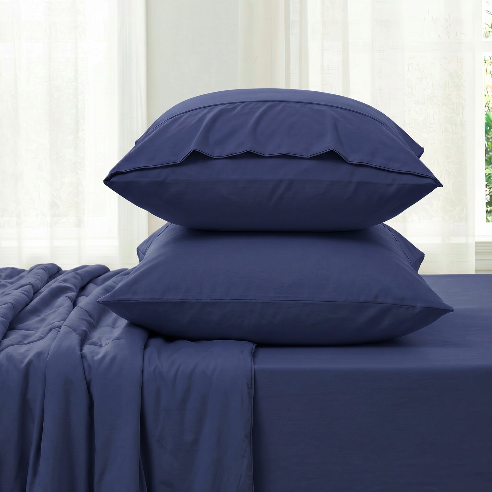 Blue bedding set with two pillowcases and fitted sheet against a white curtain background in side view.#color_Navy Blue