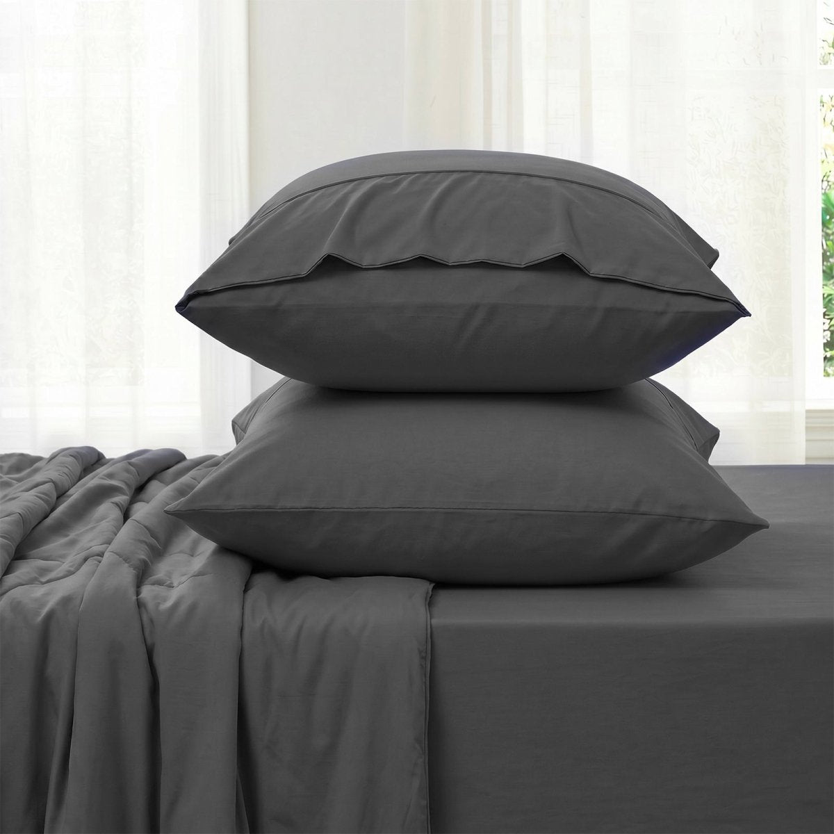 Gray bedding set with two pillowcases and fitted sheet against a white curtain background in side view.#color_Charcoal Grey