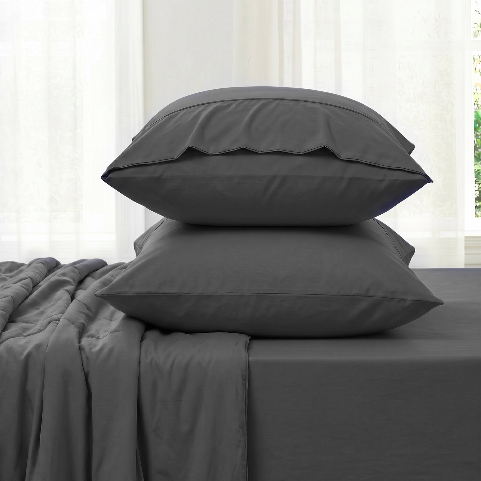 Gray bedding set with two pillowcases and fitted sheet against a white curtain background in side view.#color_Charcoal Grey
