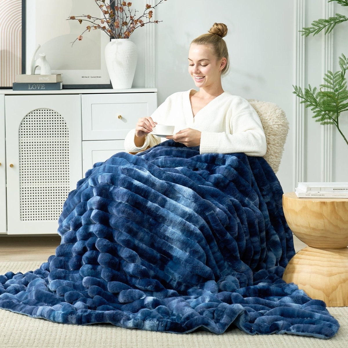 Woman sitting on a couch wrapped in a large tie-dye blue blanket in a cozy living room. #color_Tie-dye Blue