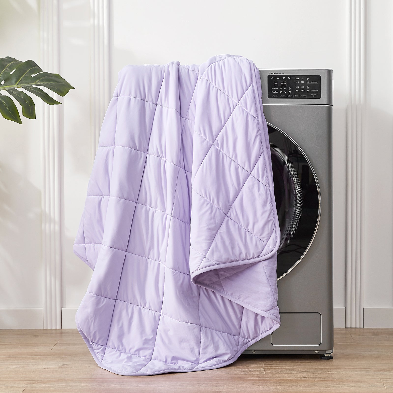 Purple quilt draped over a washing machine in a room with white walls and a plant.  #color_Purple
