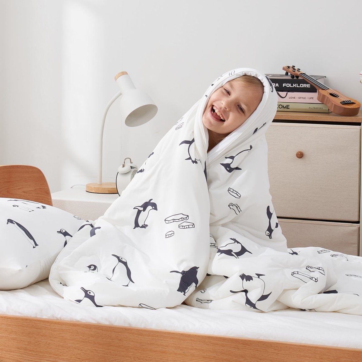 Child wrapped in a white cooling comforter with antarctic penguin prints, sitting on a bed.#color_Antarctic Penguin