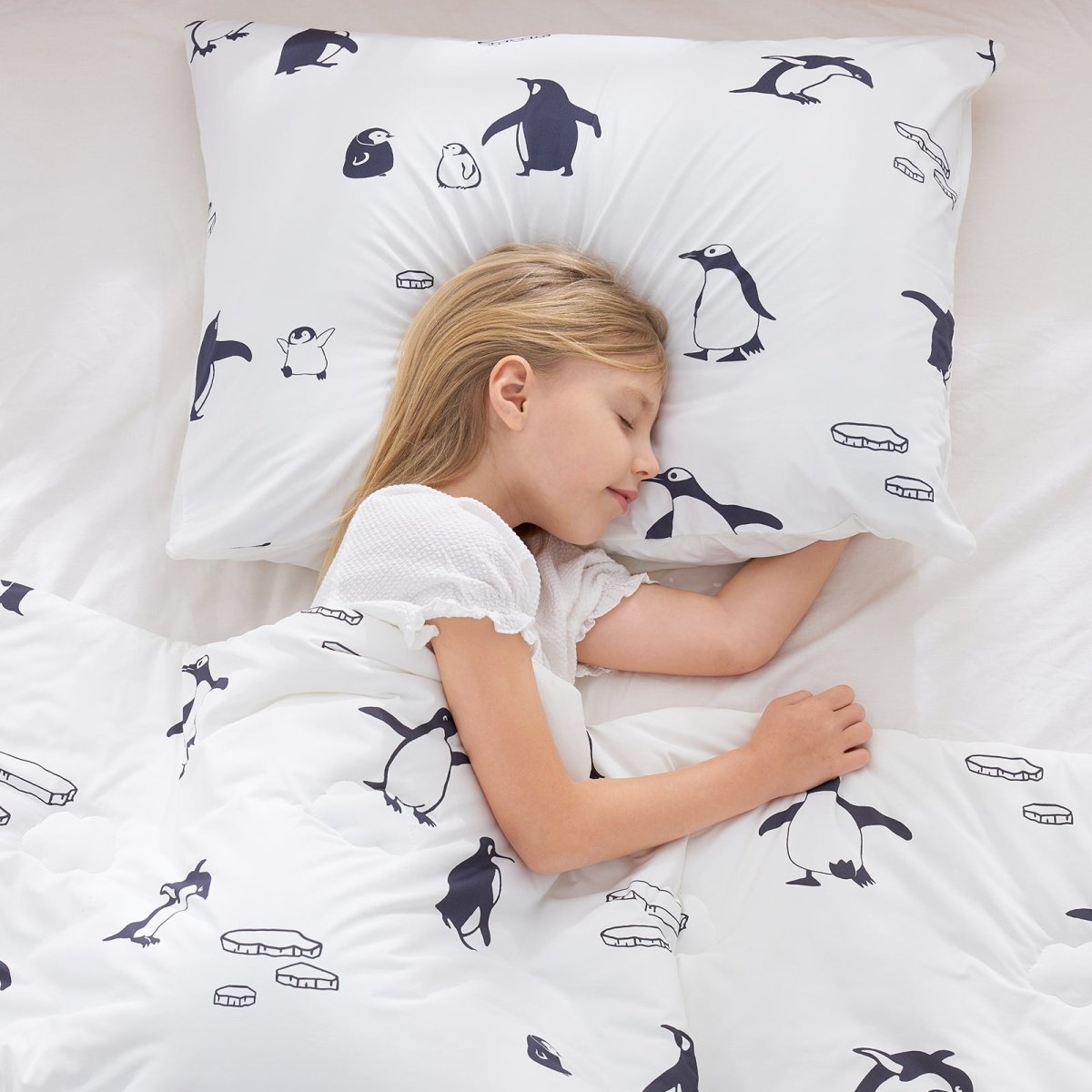 Child sleeping under a white comforter and pillow with penguin patterns.#color_Antarctic Penguin